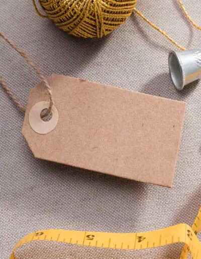Blank kraft paper clothing tag surrounded by tailoring tools including a measuring tape, safety pins, gold thread, and a thimble, symbolizing private label apparel branding and production.
