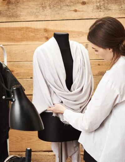Fashion designer styling a draped garment on mannequin inside a modern wooden workspace – Ibex Fashion private label clothing manufacturer.